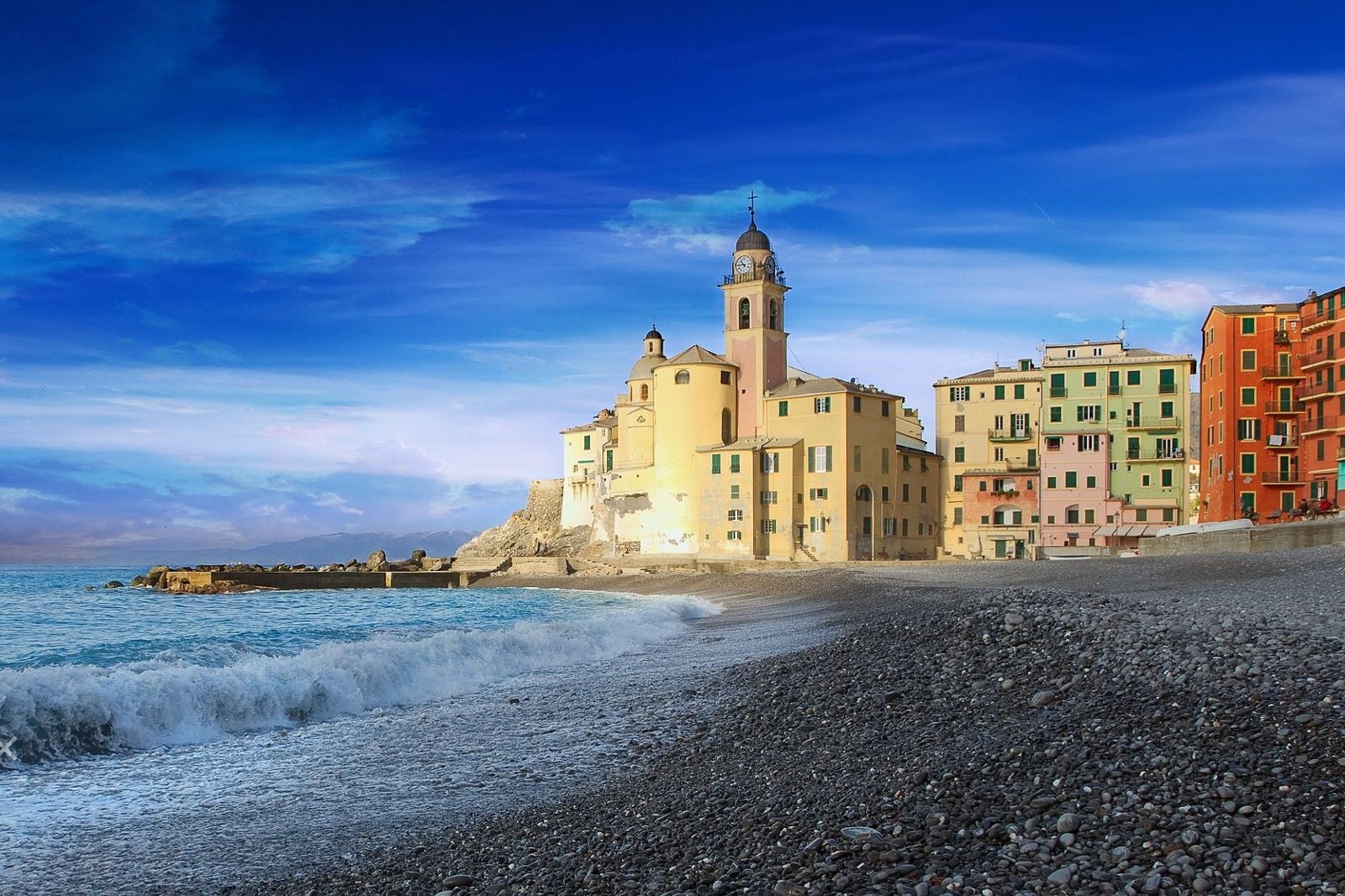Colorful buildings line the coast of a Mediterranean village, with waves gently lapping at the pebbled beach under a blue sky.